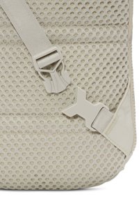 Beige ryggsäck med ett meshmönster, justerbara remmar och en kraftig plastspänne. Texturerad tyg förbättrar grepp och ventilation.