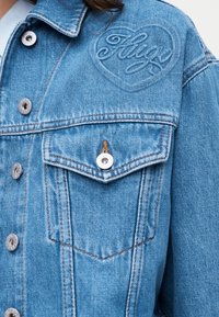 Ανοιχτό γαλάζιο τζιν μπουφάν με ασημένια κουμπιά, τσέπη στο στήθος και ανάγλυφο "Hugo" μέσα σε καρδιά στην πάνω αριστερή πλευρά.