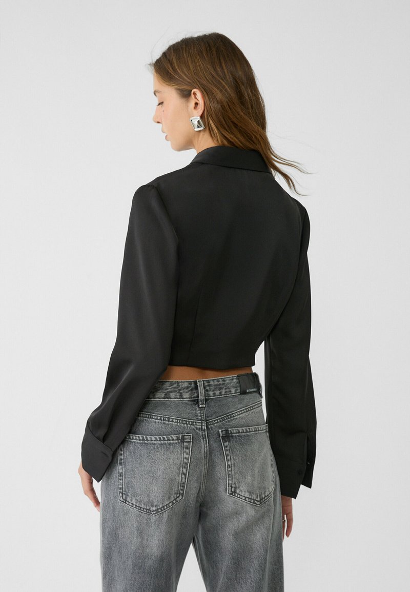 Blouse noire courte à manches longues et col, associée à un jean en denim gris délavé taille haute, présentant un design minimaliste.