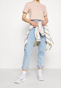 Lyhyeksi leikattu vaaleanpunainen t-paita, jossa on brodeerattu logo, yhdistettynä vaaleansinisiin farkkuihin, valkoisiin lenkkareihin ja valko-beige-raitaiseen takkiin, joka on sidottu vyötärölle.