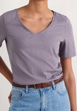 Person trägt ein hellviolettes T-Shirt mit V-Ausschnitt und kurzen Ärmeln, das in hellblaue Jeans mit braunem geflochtenem Gürtel gesteckt ist.