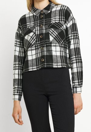 Vrouw draagt een zwart-wit geruite jas met knopen, voorvakjes op de borst en lange mouwen, gecombineerd met een zwarte broek.