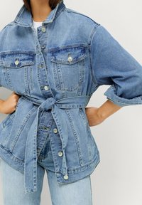 Ljusblå denimjacka med bälte i midjan, två bröstfickor, knappstängning och uppvikta ärmar. Har en strukturerad silhuett.