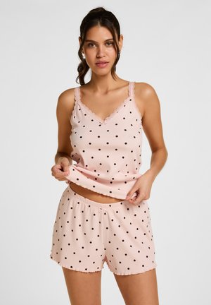 Vrouw die een roze mouwloos top met stippen en bijpassende shorts draagt, poseert tegen een effen lichte achtergrond.