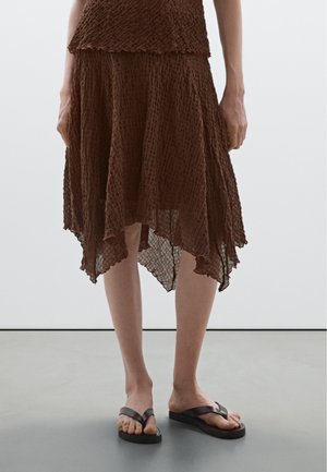 Woman wearing a brown textured handkerchief-hem skirt and black platform flip-flops standing on a gray floor.