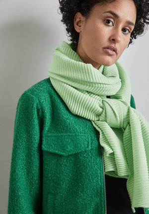 Grüne strukturierte Jacke mit aufgesetzten Taschen und einem hellgrünen gerippten Schal, der um den Hals drapiert ist, vor einem neutralen Hintergrund.