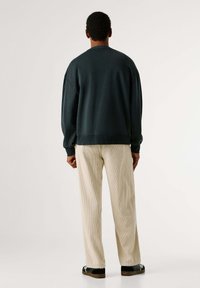 Felpa verde scuro con vestibilità rilassata, polsini a costine e collo rotondo; abbinata a pantaloni in corduroy beige chiaro e sneakers nere.