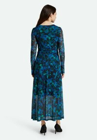 Langärmliges Blumenkleid in Blau und Grün, mit tailliertem Oberteil und transparentem Rock. Hergestellt aus leichtem, fließendem Stoff. Schwarze Pumps.