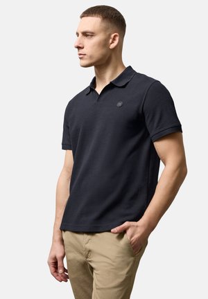 Ung mand iført en navyblå polo og beige bukser, stående med den ene hånd i lommen og kiggede til siden mod en ensfarvet baggrund.