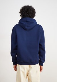 Marineblauwe hoodie van zachte stof, met een grote capuchon, ribbelmanchetten en een ontspannen pasvorm. Gedragen over lichte broek.