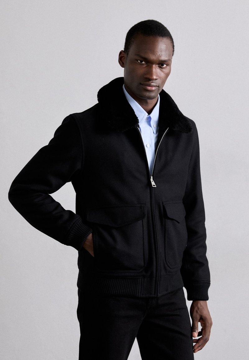 Svart zip-up jacka med krage i konstläder, ribbade ärmslut och två framfickor. Bärs över en ljusblå skjorta. Slät tygtextur.