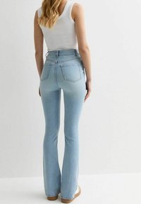 Žena stojí, má na sobě světle modré džínové kalhoty s vysokým pasem a bílý top bez rukávů, na jednoduchém bílém pozadí.