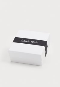 Vit rektangulär låda med ett svart horisontellt band med "Calvin Klein" i vit text. Slät, matt yta. Stängd, inga synliga innehåll.