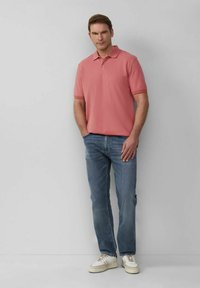 Hellrosa Polo-Shirt mit kurzen Ärmeln, kombiniert mit hellblauen Jeans und weißen Sneakers, vor einem neutralen Hintergrund stehend.