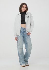 Felpa grigia con zip e logo frontale, top nero corto e jeans cargo azzurri chiari con tasche laterali. Calzature casual con suole beige.