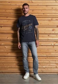 Námořnická modrá bavlněná tričko s grafickým textem, v kombinaci s lehkými džíny a bílými teniskami. Model stojí před dřevěným pozadím.