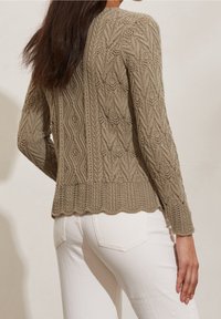 Maglione a maglia color beige con intricati motivi a onda e foglia, orlo ondulato e maniche lunghe. Indossato con jeans bianchi, che mettono in mostra texture e design.
