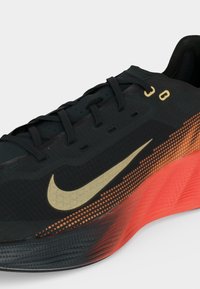 Μαύρο αθλητικό παπούτσι με χρυσό λογότυπο Nike, μαύρα κορδόνια και κόκκινη υφή στην σόλα που γίνεται μαύρη κοντά στα δάχτυλα.