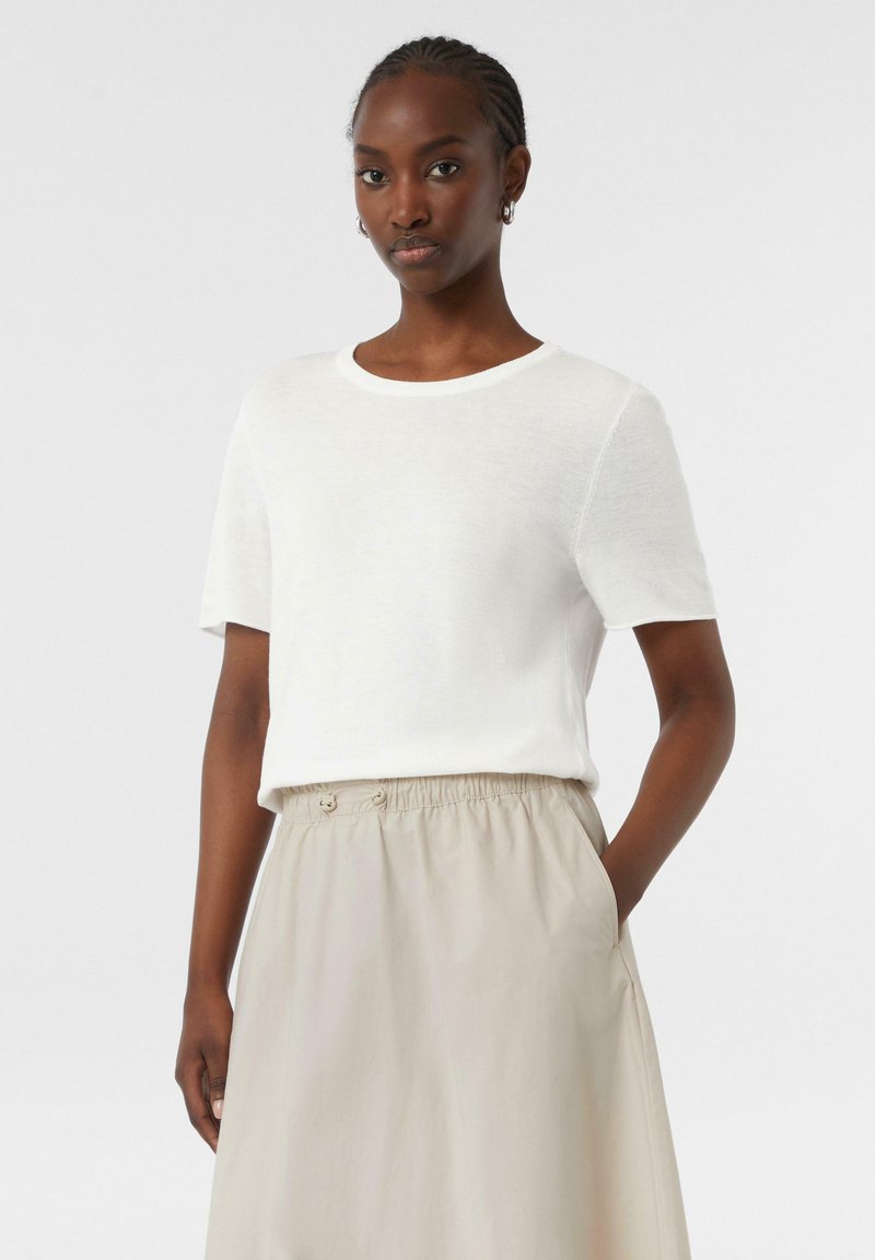 Witte kortekorte mouwtrui met een ronde halslijn, zachte textuur, gedragen met een beige rok met elastische taille en zijzakken.