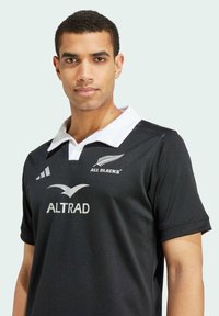 Sort rugbytrøje med hvid krave, prydet med "ALL BLACKS" logo og "ALTRAD" tekst. Glat stof med adidas tre striber på skulderen.