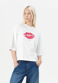Weißes T-Shirt mit kurzen Ärmeln, das ein rotes Lippenmotiv in der Mitte zeigt; lockere Passform, weicher Textur, mit rundem Ausschnitt und verkürzter Länge.
