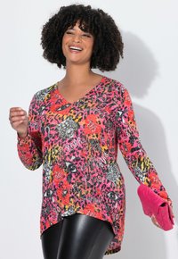 Femme souriante aux cheveux bouclés portant un haut à manches longues en imprimé animal coloré et un pantalon en cuir noir, tenant une pochette matelassée rose vif.