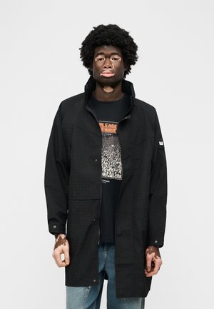 Άνδρας με λευκά στίγματα στο δέρμα (βιτιλίγκο), φοράει μαύρο σακάκι πάνω από ένα γραφικό μπλουζάκι και μπλε τζιν, στέκεται μπροστά σε ένα απλό φωτεινό φόντο.