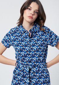 Vestito camicia azzurro a maniche corte con un motivo di uccelli stilizzati, caratterizzato da una chiusura frontale con bottoni e una cintura annodata in vita. Tessuto dalla superficie liscia.