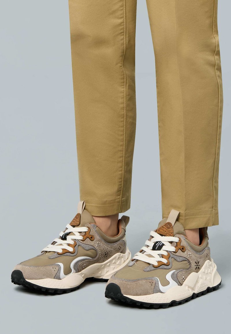 Scarpe da ginnastica beige con materiali misti in suede e rete, dettagli bianchi, suola nera e un'etichetta sul tallone texturizzata. Abbinate a pantaloni beige.