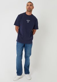 Hombre de pie con las manos detrás de la espalda, vistiendo una camiseta azul marino con la palabra "Provence", jeans azules y zapatillas blancas sobre un fondo blanco.