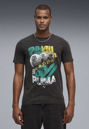 Mann trägt ein schwarzes Puma T-Shirt mit dem Schriftzug „Brazil“, drei Fußbällen, fünf Sternen und grünen sowie gelben grafischen Elementen.