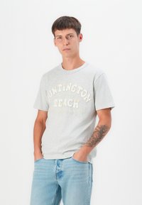 Pilkas medvilnės T-shirt su trumpomis rankovėmis, ant krūtinės iškilmingai užrašyta "Huntington Beach" šviesiomis raidėmis. Prigludęs dizainas.