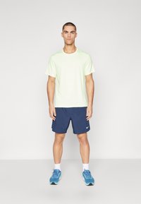 T-shirt atletico verde chiaro a maniche corte, pantaloni corti blu e scarpe da corsa blu. La t-shirt presenta un logo bianco sul petto.