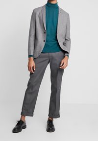 Man med grå kavaj, teal polotröja, mörkgrå byxor med fåll och svarta snörskor, står mot en neutral bakgrund.