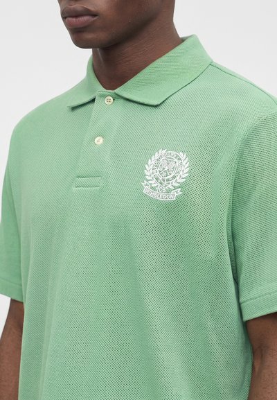 Άνδρας που φοράει ανοιχτό πράσινο polo Wimbledon με λευκό κεντημένο λογότυπο στο αριστερό στήθος, κοντά μανίκια και κουμπωτό γιακά.