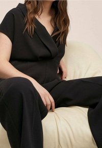 Zwarte jumpsuit met korte mouwen en een V-hals, gemaakt van een zachte stof, met een relaxte pasvorm en zichtbare naden, gestyled tegen een lichte achtergrond.