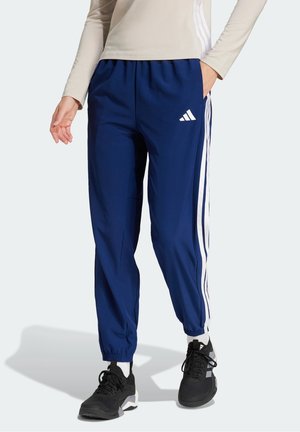 Osoba nosząca granatowe spodnie dresowe Adidas z białymi paskami i logo, beżową bluzkę z długim rękawem, czarne sneakersy i białe skarpetki.