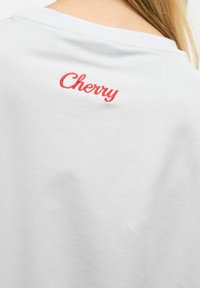 T-shirt en coton blanc avec un col rond et un texte "Cherry" brodé en rouge dans le dos. Texture lisse, design décontracté.