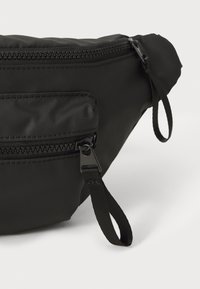 Sac banane noir fabriqué en nylon durable. Il dispose d'un compartiment principal et d'une petite poche zippée à l'avant, avec une sangle ajustable et des accents en métal.