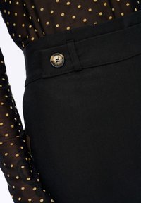 Zwarte broek met een gladde textuur, voorzien van een knoopaccent. Een doorzichtige top met stippen en lange mouwen is gedeeltelijk zichtbaar.