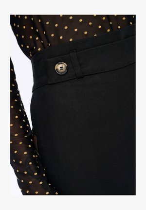 Zwarte broek met een gladde textuur, voorzien van een knoopaccent. Een doorzichtige top met stippen en lange mouwen is gedeeltelijk zichtbaar.