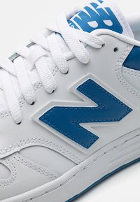 Bílé kožené tenisky s výrazným modrým logem "N", texturovanými akcenty, bílými tkaničkami a vzorovaným modrým jazykem.