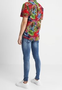 Rotes, kurzärmeliges Hemd mit Blumenmuster und grünen sowie blauen Blattmotiven, kombiniert mit blauen Skinny-Jeans und weißen Sneakern, von hinten gesehen.