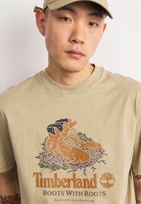 T-shirt en coton beige avec un design illustré de bottes avec des racines, dans des tons terreux et le texte "Bottes Timberland avec Racines".