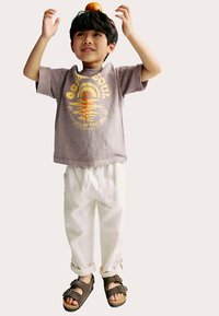 Camiseta de color gris claro con un diseño gráfico colorido, pantalones sueltos blancos y sandalias marrones. El niño sostiene una naranja por encima de su cabeza, sonriendo.