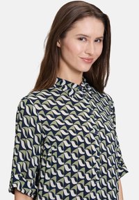 Blusa con bottoni di colore blu navy con un motivo ripetuto di onde verdi e bianche. Presenta maniche corte e un colletto. Tessuto dalla texture liscia.