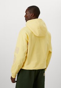 Yellow hoodie with a drawstring hood, ribbed cuffs, and back graphic. Worn with green pants, showcasing a relaxed fit and soft texture.