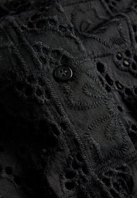 Μαύρο ύφασμα με κεντήματα λουλουδιών και σχέδια με κοψίματα, με μια μαύρη κουμπότρυπα τεσσάρων οπών ραμμένη στο κέντρο.