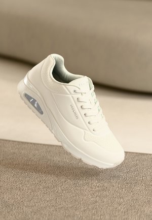 Zapatilla blanca Skechers con suela visible con cámara de aire y diseño con cordones sobre una superficie texturizada beige y fondo desenfocado.