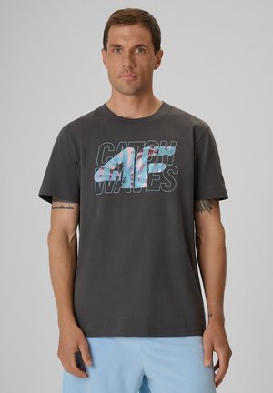 Mężczyzna z krótkimi brązowymi włosami ma na sobie ciemnoszary t-shirt z grafiką "Catch Waves" i jasnoniebieskie szorty, stojąc na tle jednolitego jasnego tła.